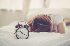 Kobieta leży w łóżku, przed nią budzik wskazujący godzinę 10:00. To nadrabianie snu w weekendy