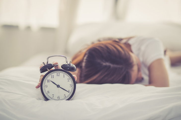 Kobieta leży w łóżku, przed nią budzik wskazujący godzinę 10:00. To nadrabianie snu w weekendy